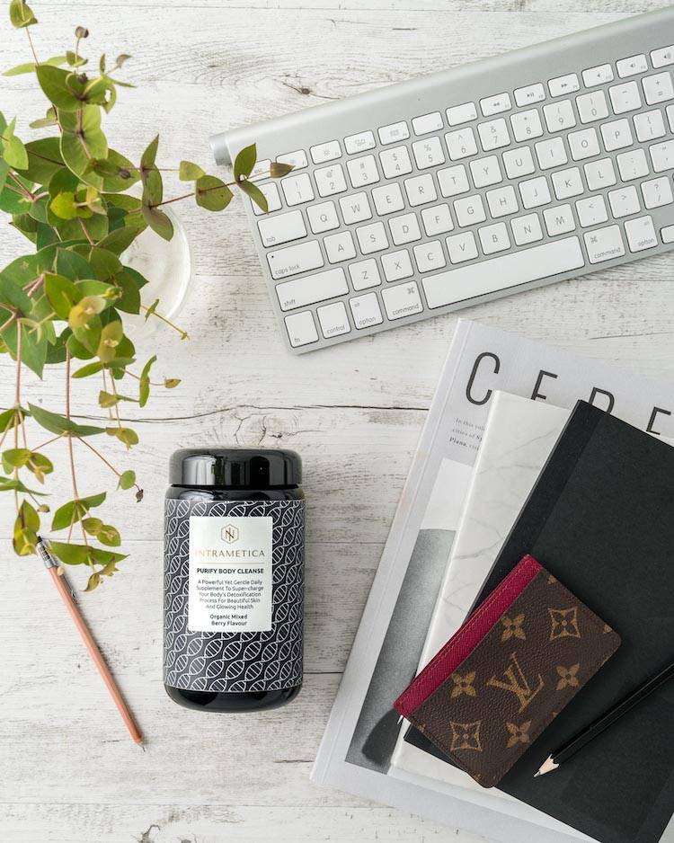 A modern workspace featuring a white keyboard, magazines, a potted green plant, a bottle of Intrametica beauty supplements, and a wallet with a fashionable pattern, all arranged on a light wooden