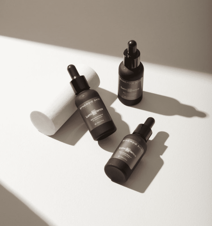 Three dark amber Synergie Skin glass bottles with droppers, labeled with white text, are arranged neatly on a light surface under natural sunlight, casting soft shadows.