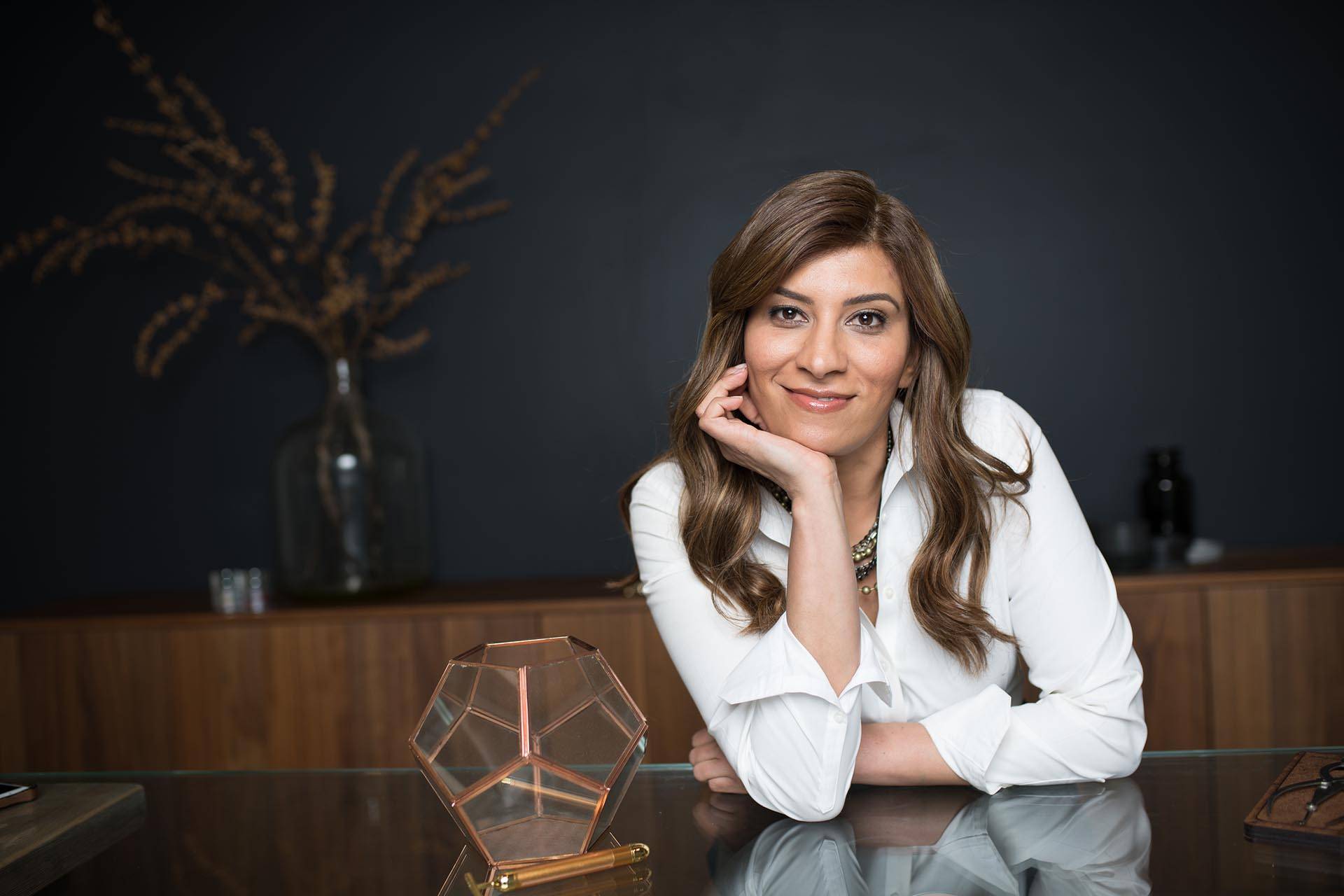 A woman in a white blouse sits at a desk, smiling at the camera with her chin resting on her hand. A geometric glass object and a vase with branches are on the table, alongside broch