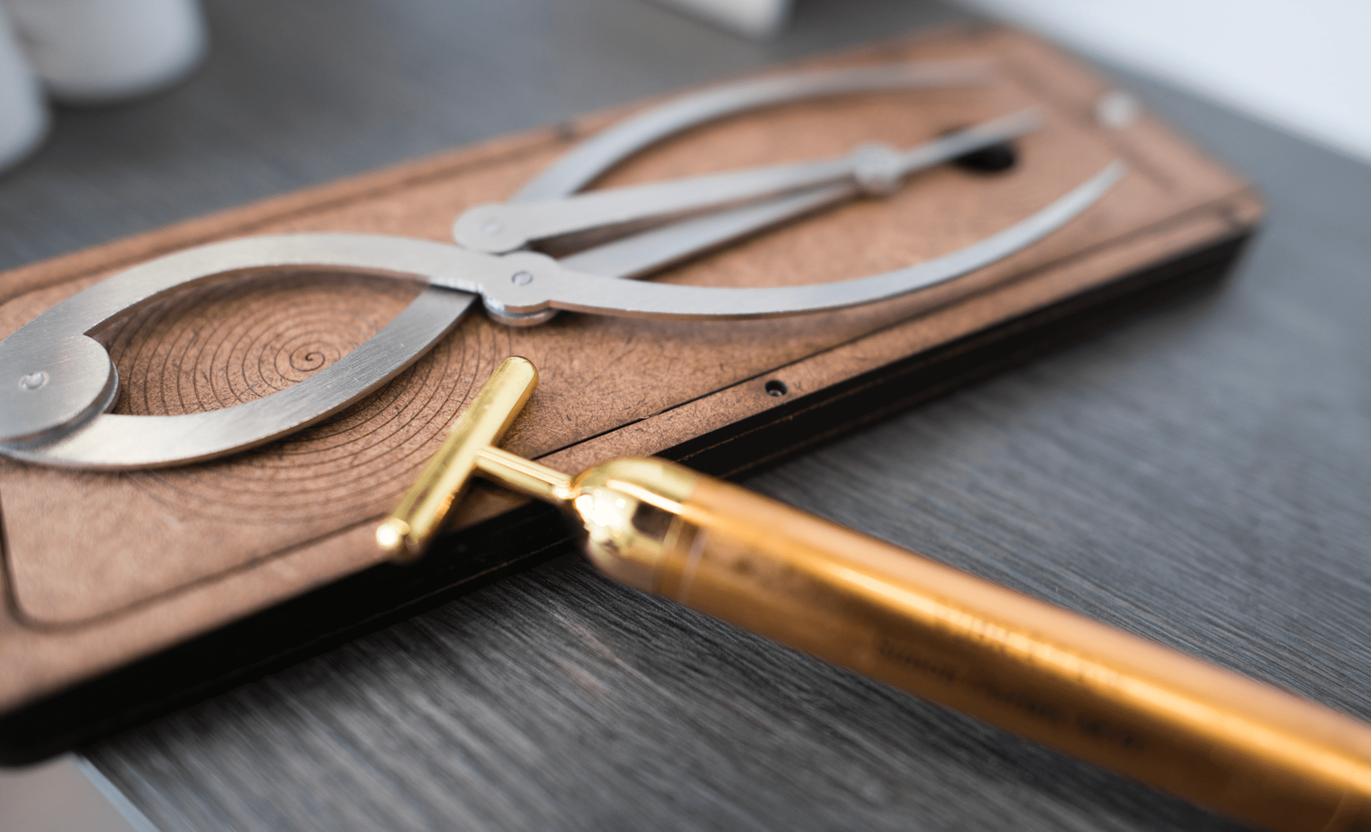 A close-up image of a wooden drawing board with a pair of large compasses and a golden mechanical pencil resting on it. The focus is sharp on the metallic elements of the tools, portraying them as precise measuring and accuracy in treatment.