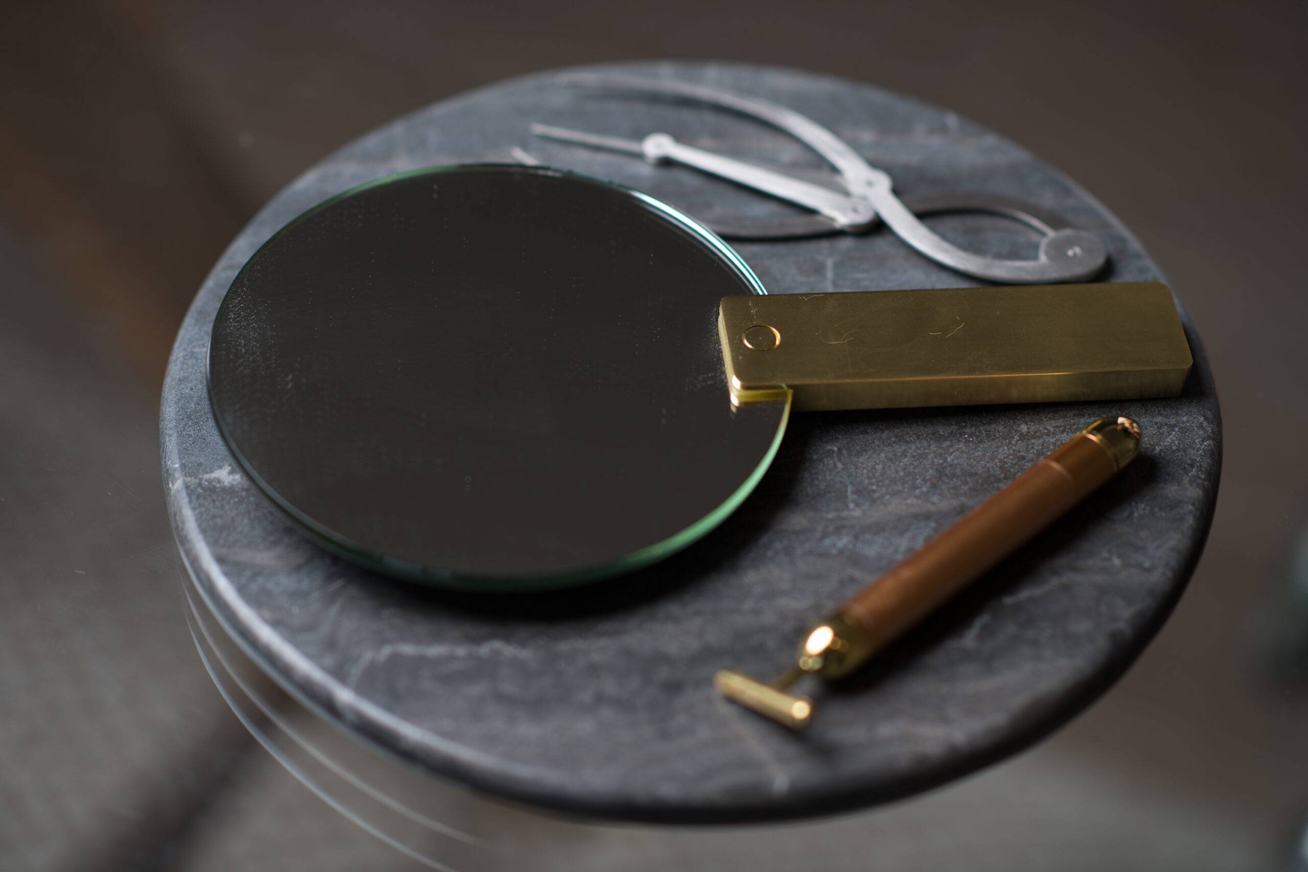 A magnifying glass with a brass handle lies on a gray stone tray next to white scissors and a brass letter opener, all meticulously arranged for revision on a smooth surface.