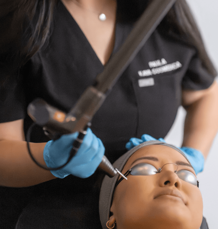 A close-up view of a woman receiving a Spectra Laser treatment, focusing on the forehead area, administered by a professional wearing a black uniform and blue gloves. The client is reclined and wearing protective
