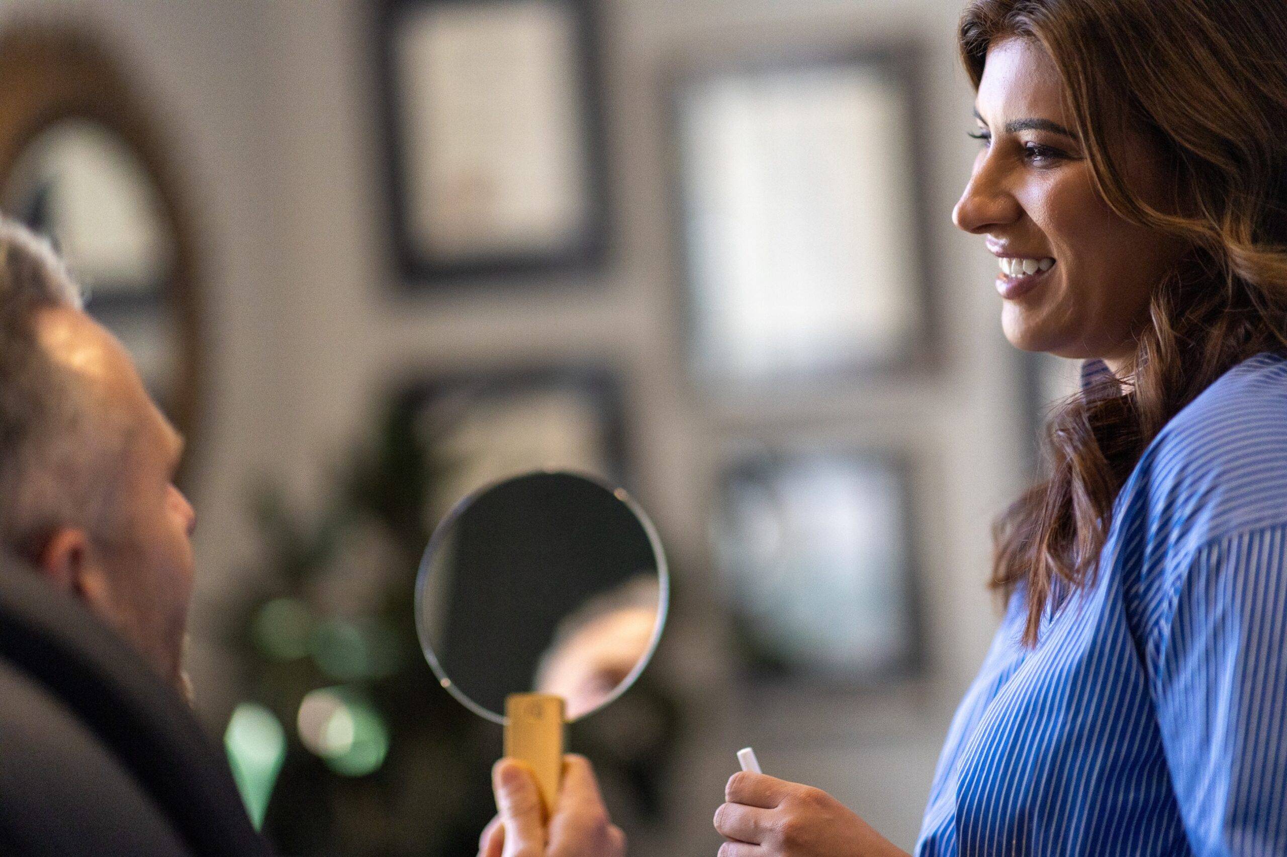 A woman with brown hair, smiling, holds a mirror and lipstick, talking to an older man whose reflection is seen in the mirror, in a warmly decorated room offering cosmetic treatments.