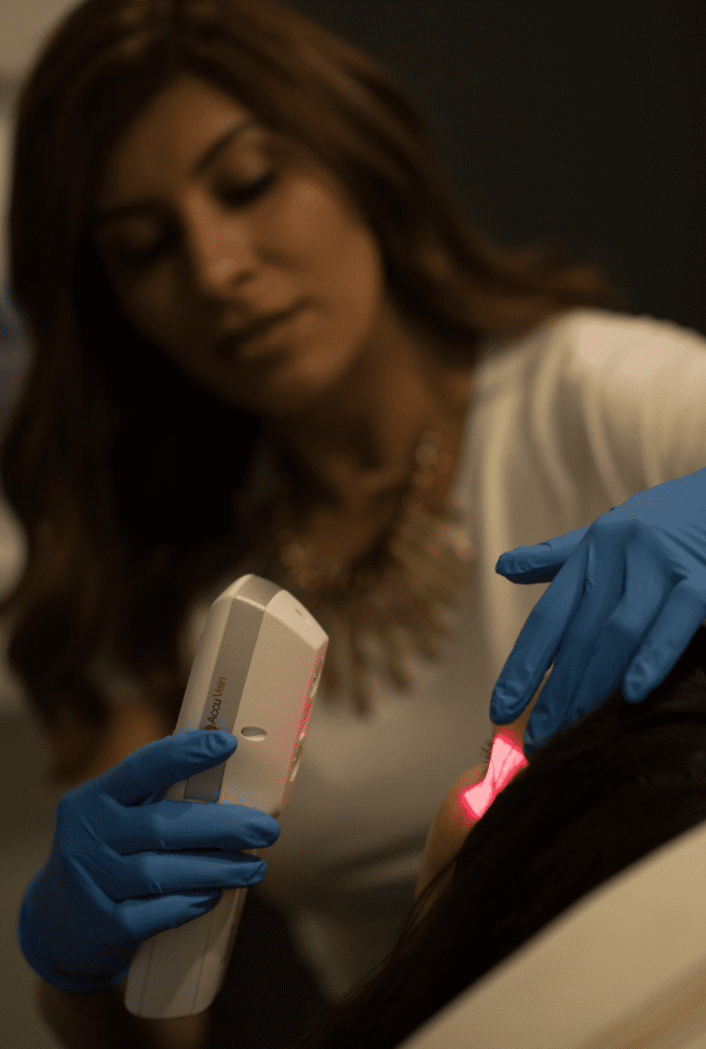 A dermatologist in blue gloves uses a handheld laser device on the scalp of a female patient to treat hair loss. The patient, wearing a white shirt, sits calmly during the procedure, which also includes