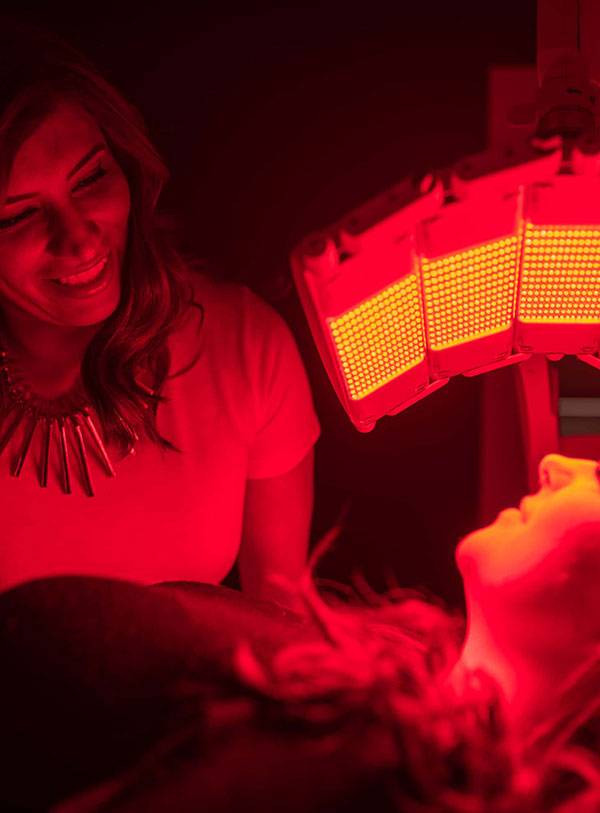 A woman smiles while administering Omnilux LED therapy to another woman lying down, bathed in red light from a panel above her face.