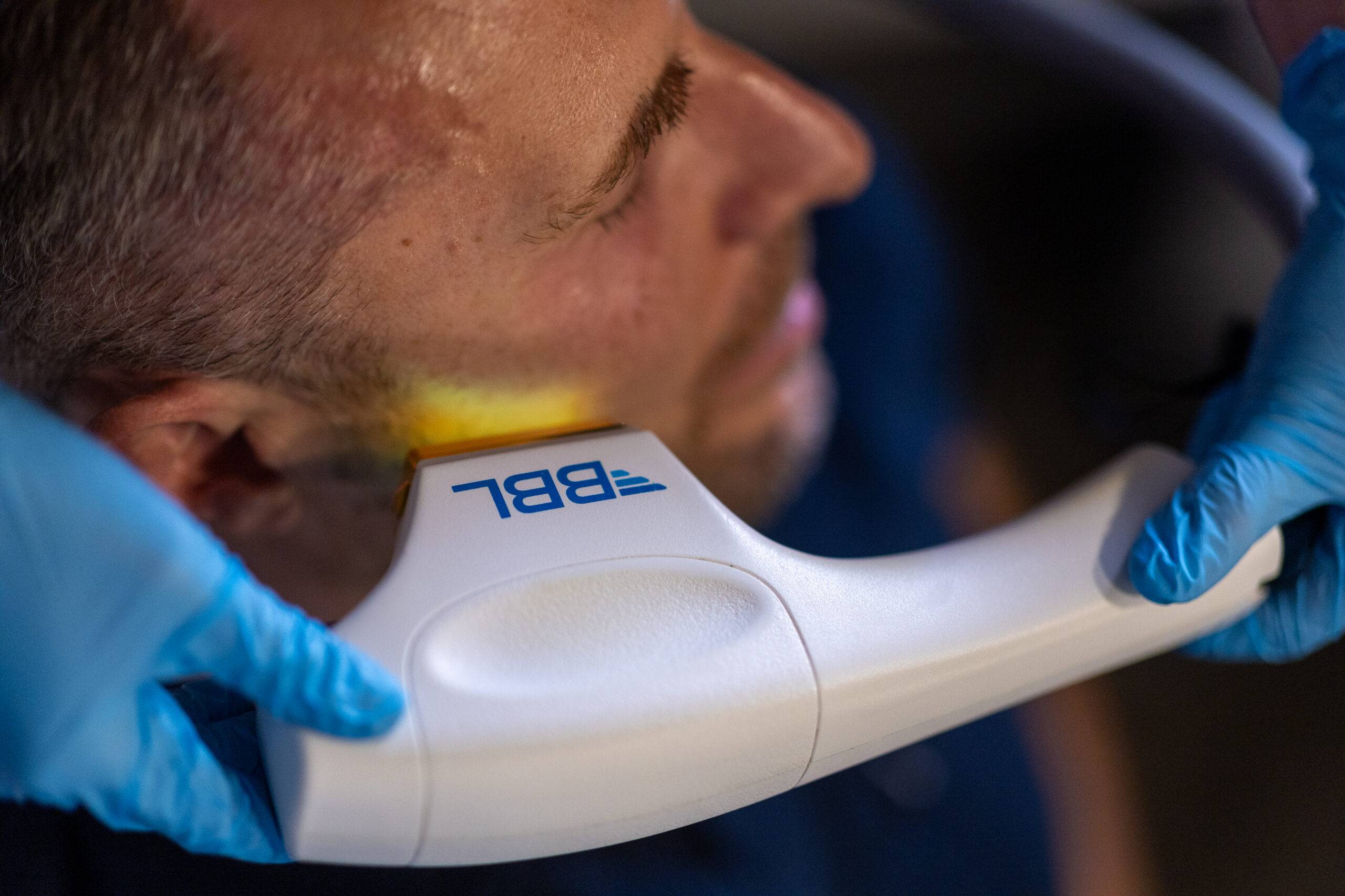 A close-up of a man undergoing a dental x-ray, with a dental assistant wearing blue gloves holding the BroadBand Light Therapy device under his chin. The device is labeled with the number "987".