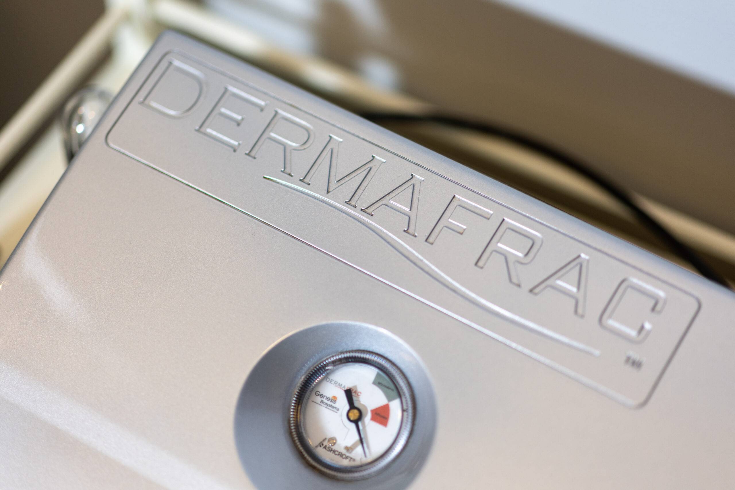 Close-up of a microneedling machine with a focus on its logo and an operational dial gauge showing a measurement in the red zone. The background is softly blurred.