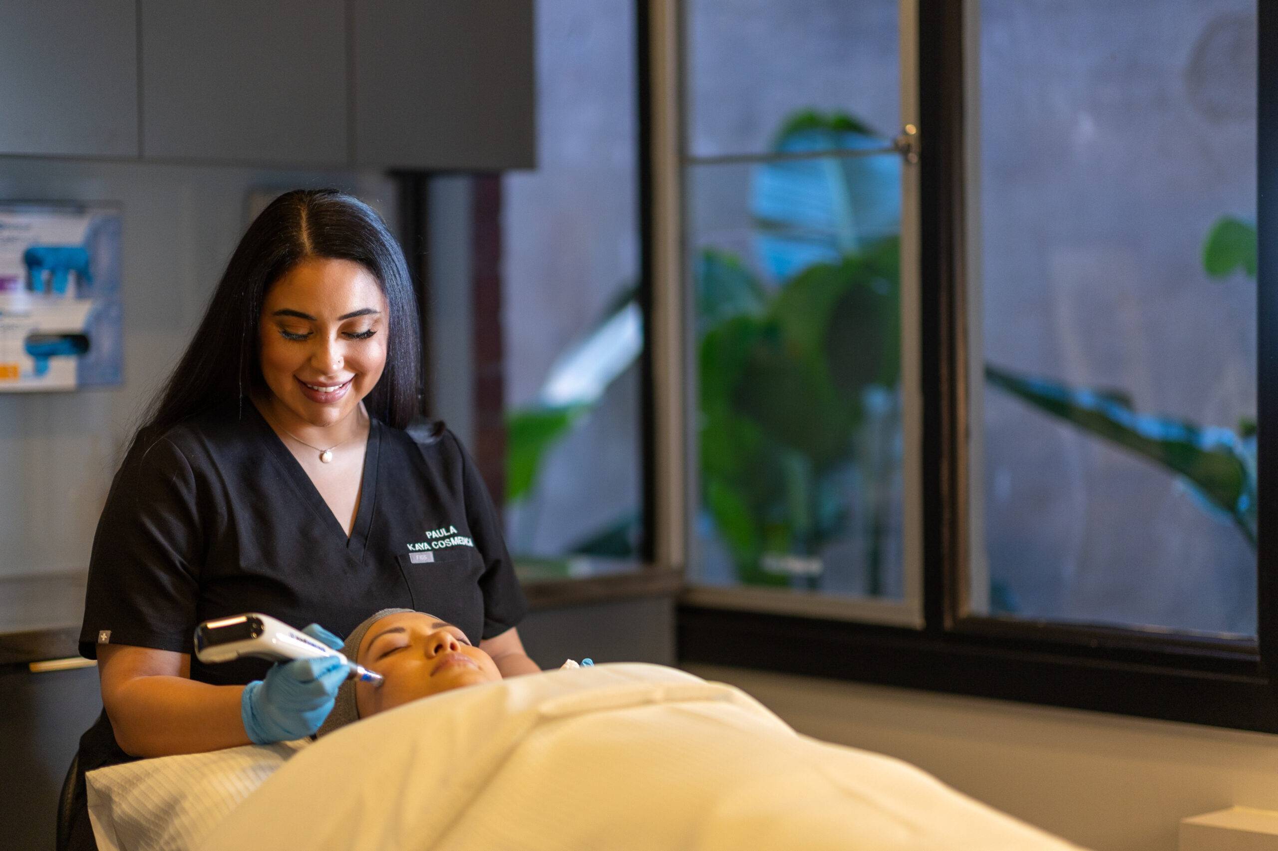 A female aesthetician in a black uniform performs a microneedling skin treatment on a reclining female client in a well-lit room with plants visible through the window.