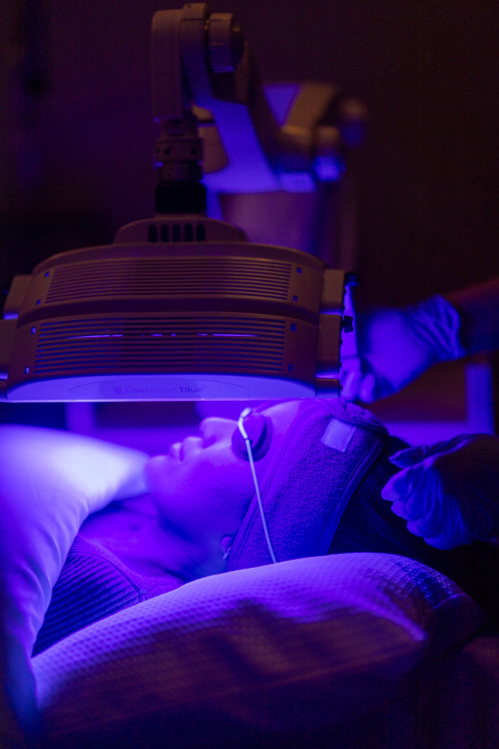 A woman undergoes a skincare treatment using the Omnilux machine emitting a blue light, in a dimly lit spa environment. She is resting on her back with eyes covered for protection.