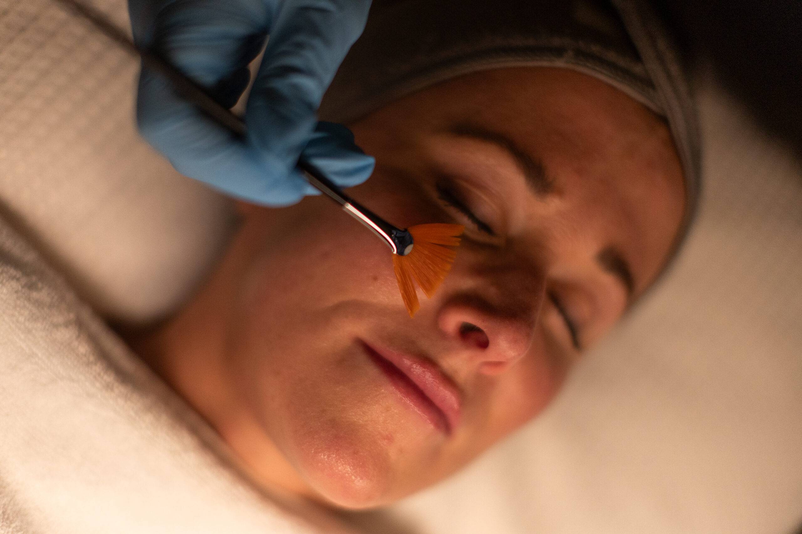 A woman receiving a skin treatment with a brush applying a product under her eyes, wearing a headband and lying down, as a person wearing blue gloves tends to her skin.