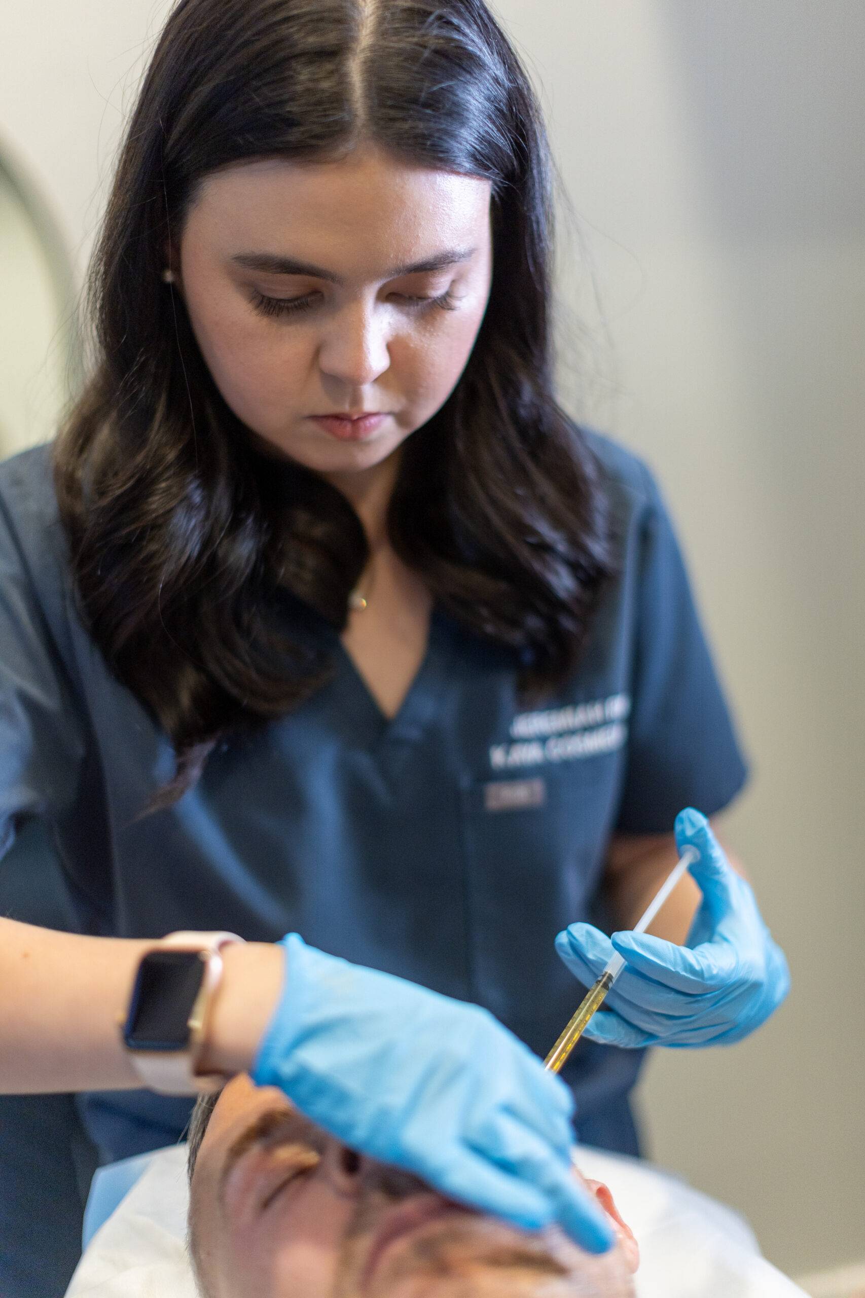 A female medical professional in blue scrubs is administering a skin treatment to a male patient lying down. She wears gloves and is focused on her task, holding a medical tool.