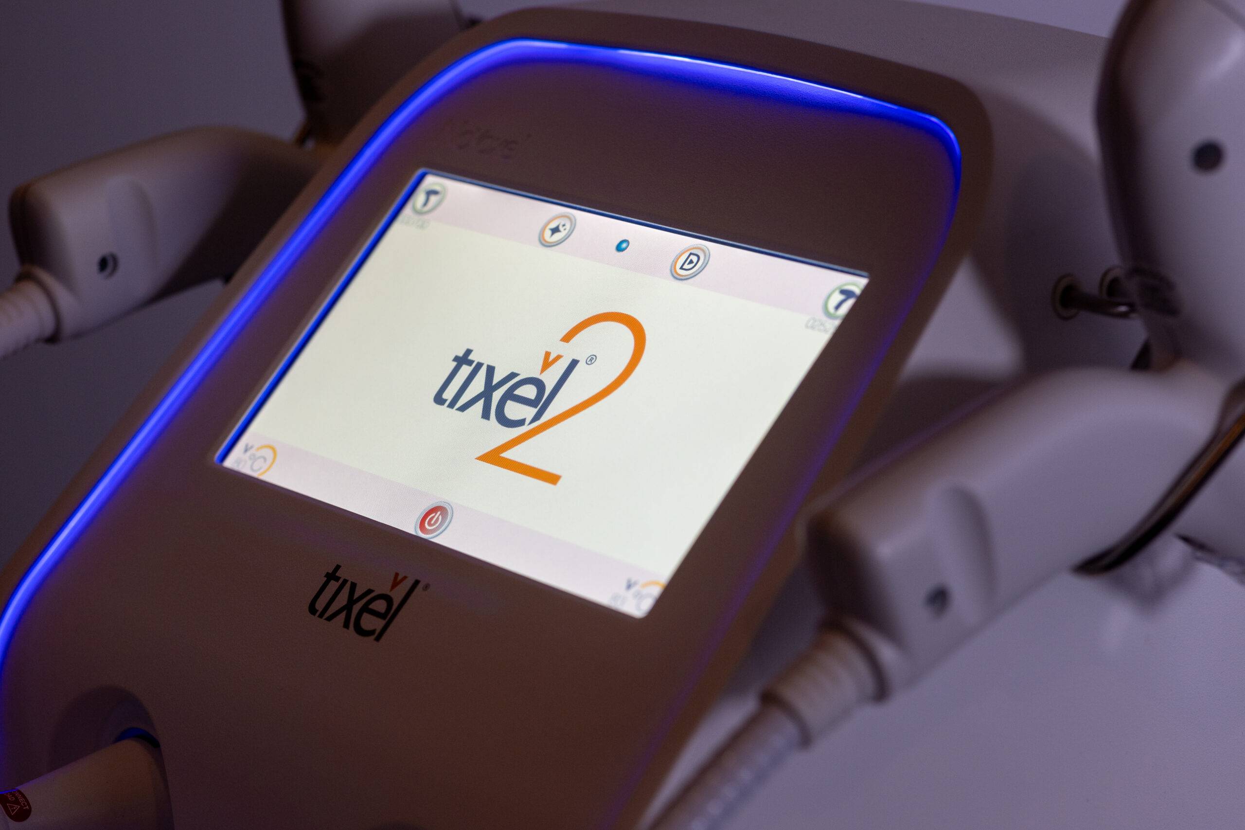 Close-up of a medical device with a screen displaying the logo "Tixel 2" highlighted by a blue light, with cables connected to its structure. The description you provided is too brief and lacks