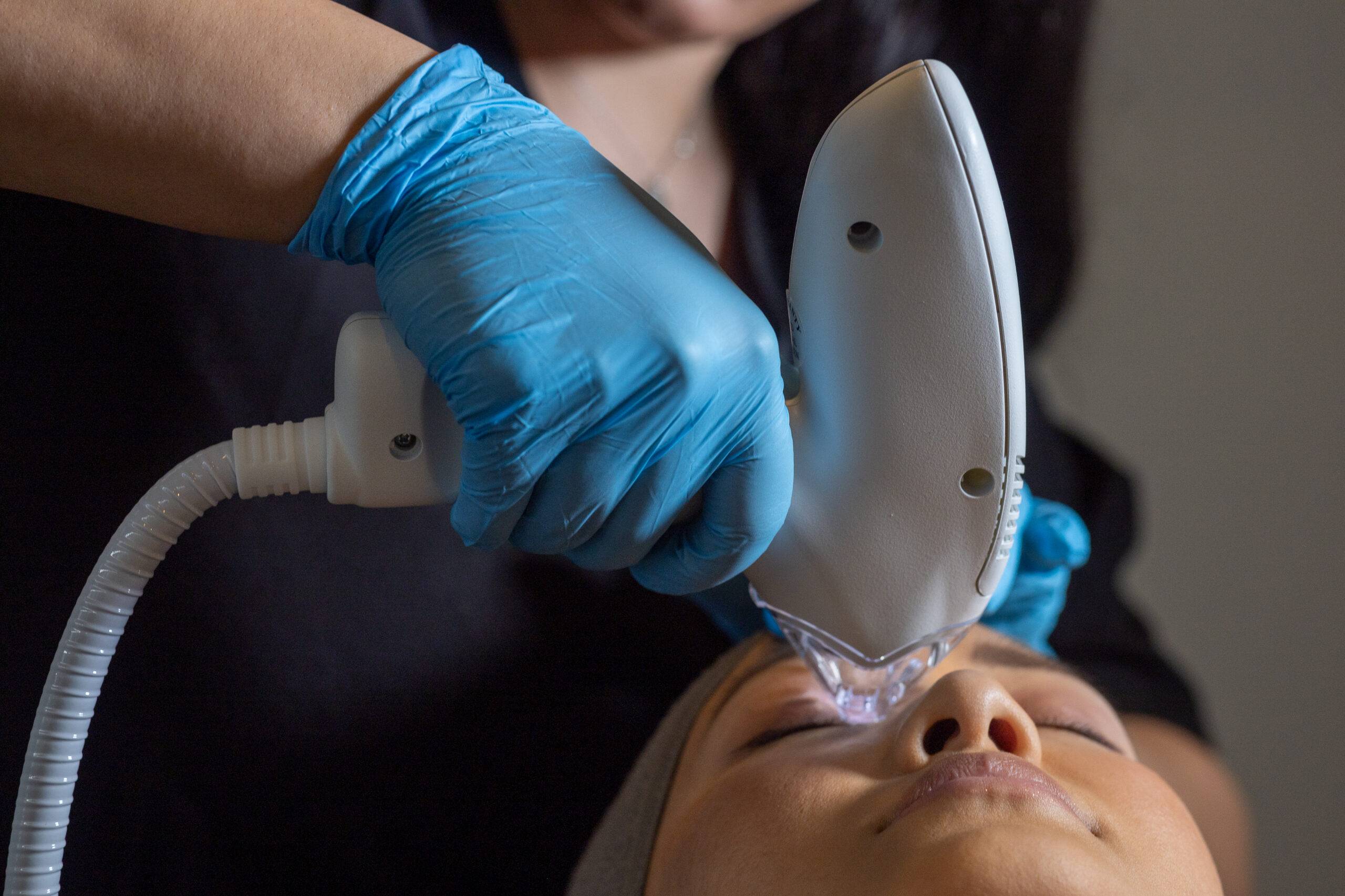 Aesthetician wearing blue gloves uses a Tixel device on the face of a reclining woman during a skincare treatment.