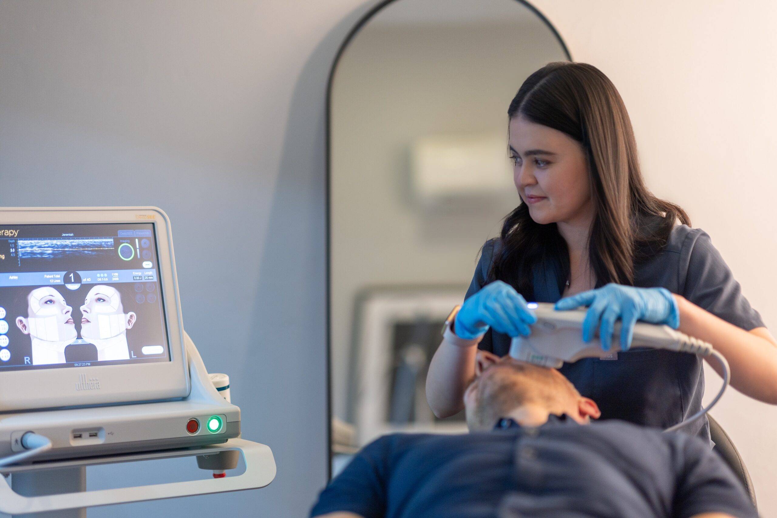 A female healthcare professional uses an Ultherapy device on a male patient's neck, viewing the results on a nearby screen in a medical office.