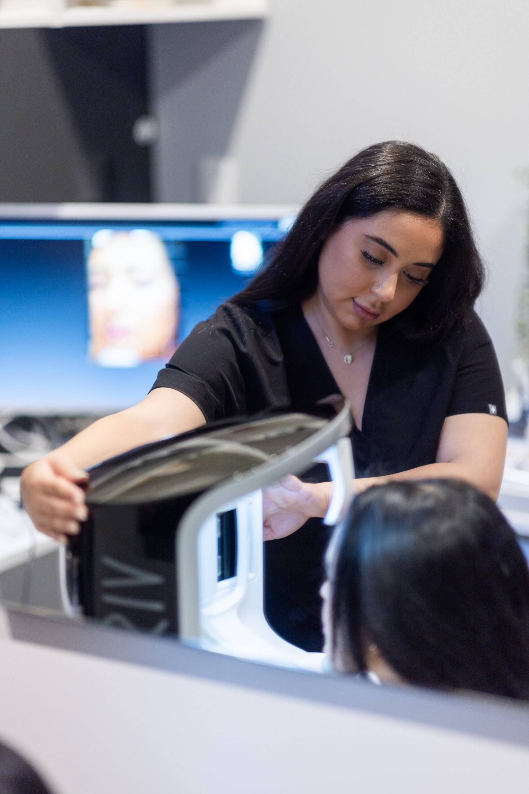 A woman with dark hair examines her reflection in a high-tech Visia mirror, adjusting equipment on a modern desktop, possibly in a beauty salon or medical clinic.
