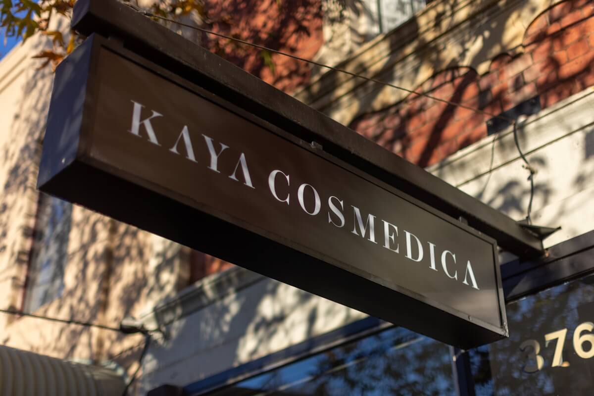 A sign reading "kaya cosmedica" hangs from a metal bracket on a sunny day, with a blurred red brick building in the background.