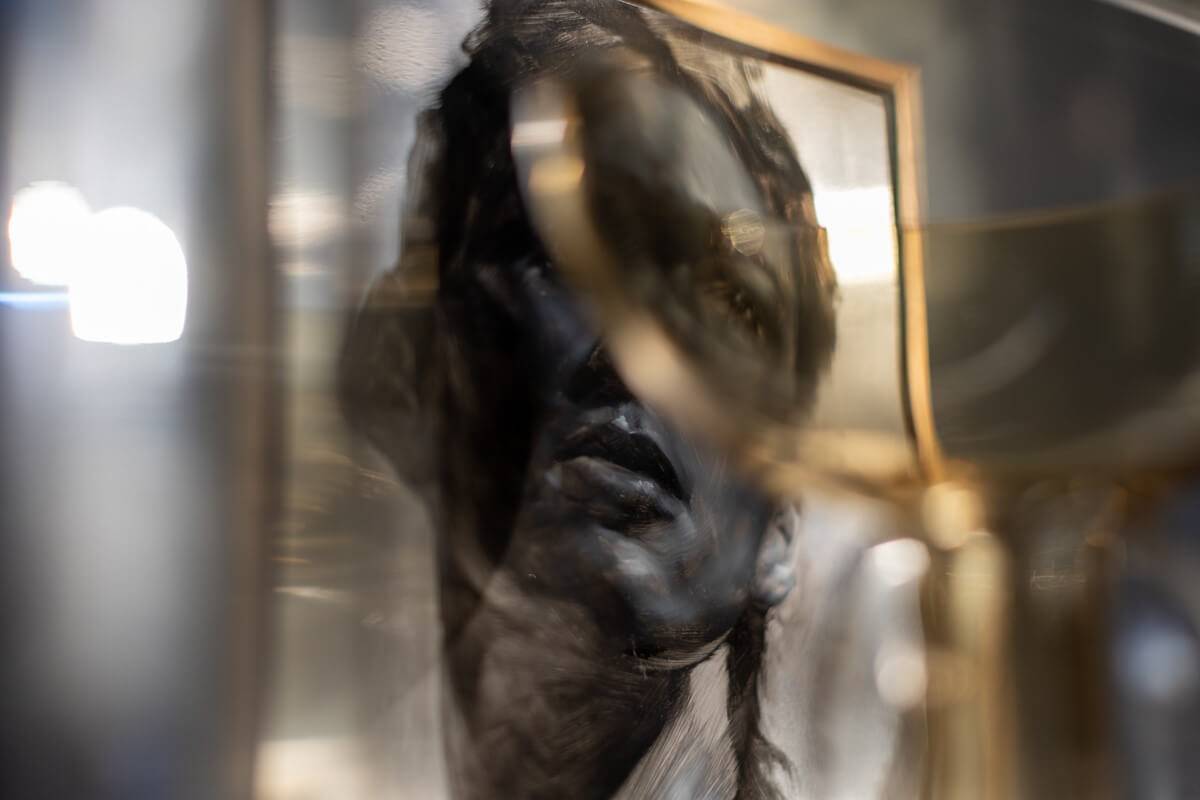 A blurred sculpture of a human face displayed behind a glass pane, with reflections and distortions adding an abstract quality to the image.