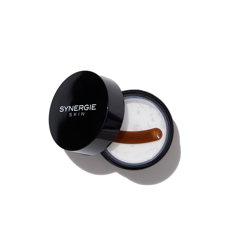 An open jar of synergie skin moisturizer with a small spatula on a white background. the jar is black and the cream is textured white.
