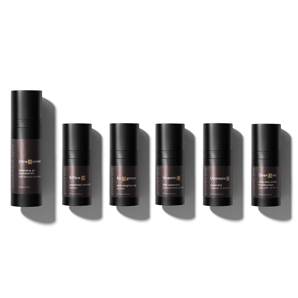 Six skincare products from the Skin Brightening Kit in black cylindrical containers, arranged in a row on a light gray background, casting shadows to the right. Labels on containers display various skincare ingredients.