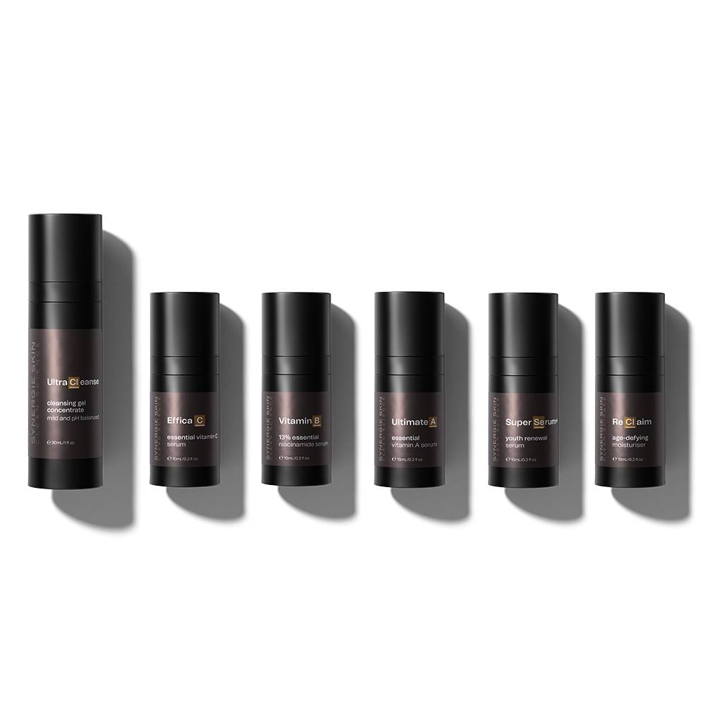 Six cylindrical Synergie skincare product containers from the Ultimate Anti-Ageing Kit in varying shades of brown with white labels lie in a row on a light grey background, exhibiting different product names and descriptions.