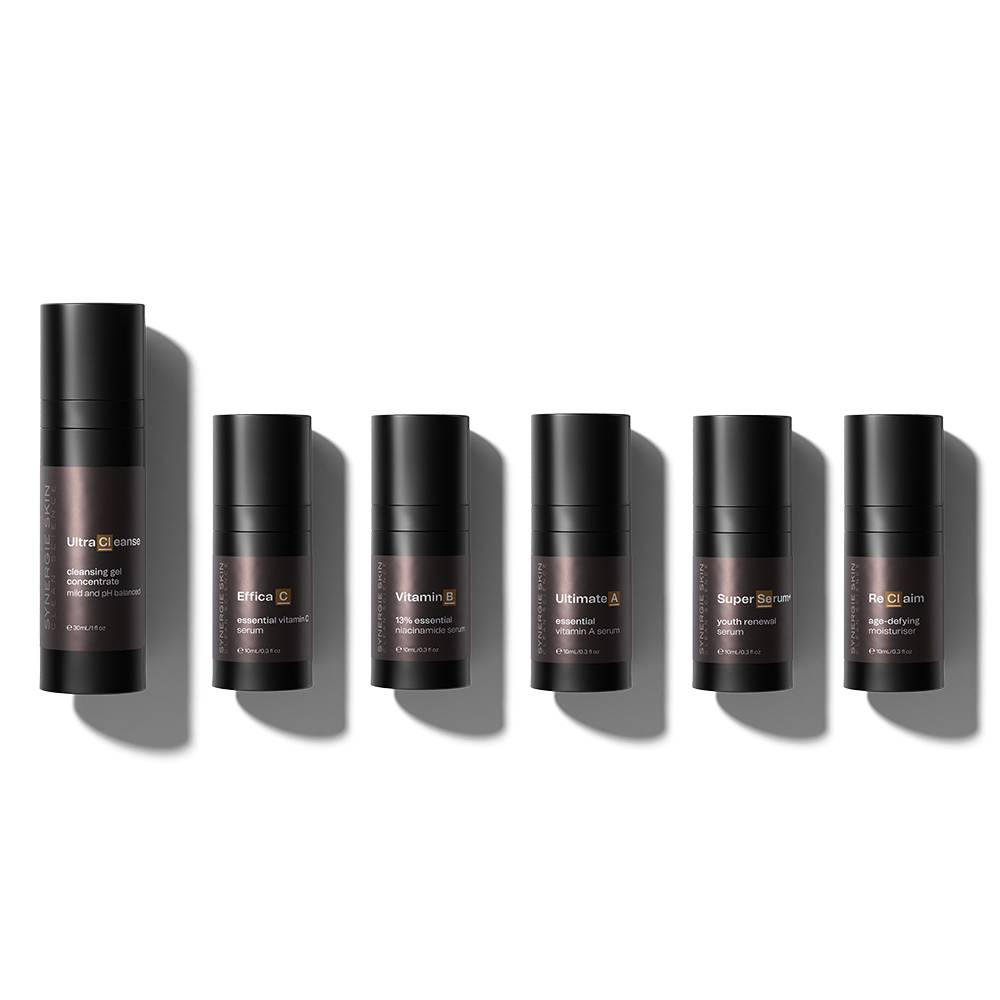Six cylindrical Synergie skincare product containers from the Ultimate Anti-Ageing Kit in varying shades of brown with white labels lie in a row on a light grey background, exhibiting different product names and descriptions.