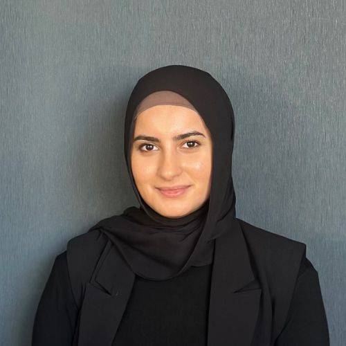 A woman wearing a black hijab with a neutral expression poses for a team portrait against a grey background. She has a slight smile and is looking directly at the camera.