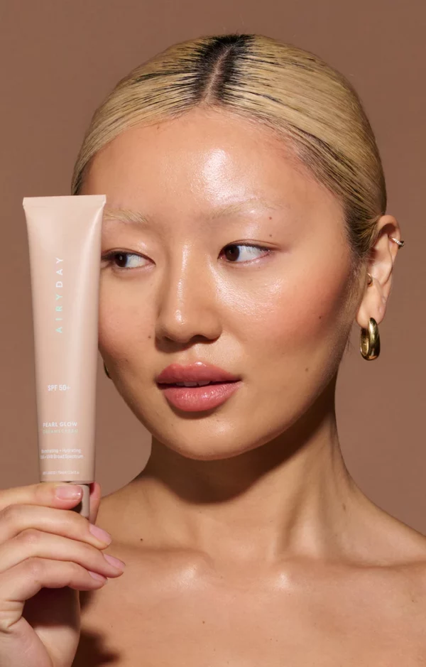 A light-skinned person with blonde hair pulled back holds a tube of Pearl Glow SPF50+ Dreamscreen 75ml. The solid brown background highlights their gold hoop earrings as they look to the side.