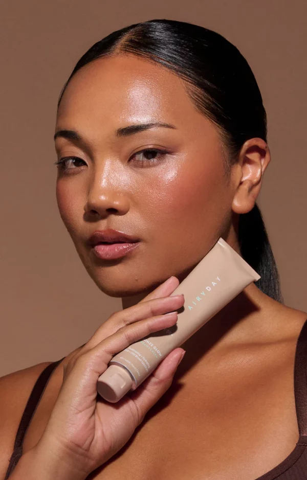 A person with smooth, glowing skin holds a 75ml tube of Bronze Glow SPF50+ Dreamscreen near their face. They have slicked-back hair and a neutral expression, set against a warm solid background.