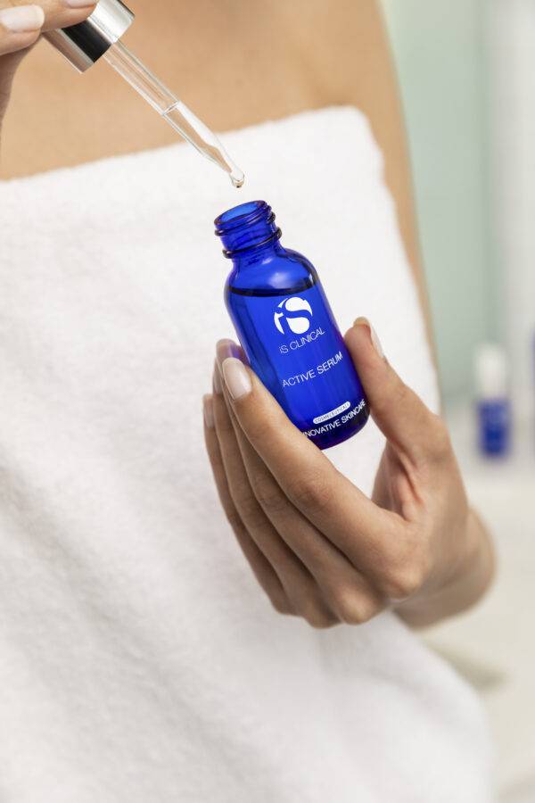 A person in a white towel holds a 30ml blue bottle labeled "Active Serum 30ml" and dispenses serum from the dropper above the bottle.