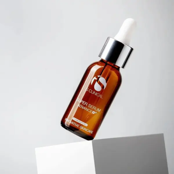A brown dropper bottle labeled “Super Serum Advance+ 15ml” is balanced on the edge of a white block against a plain light gray background.