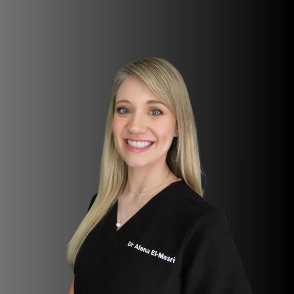 A woman with long blonde hair smiles at the camera. She is wearing a black top embroidered with "Dr Alana El-Masri" and stands in front of a gradient gray-to-black background.