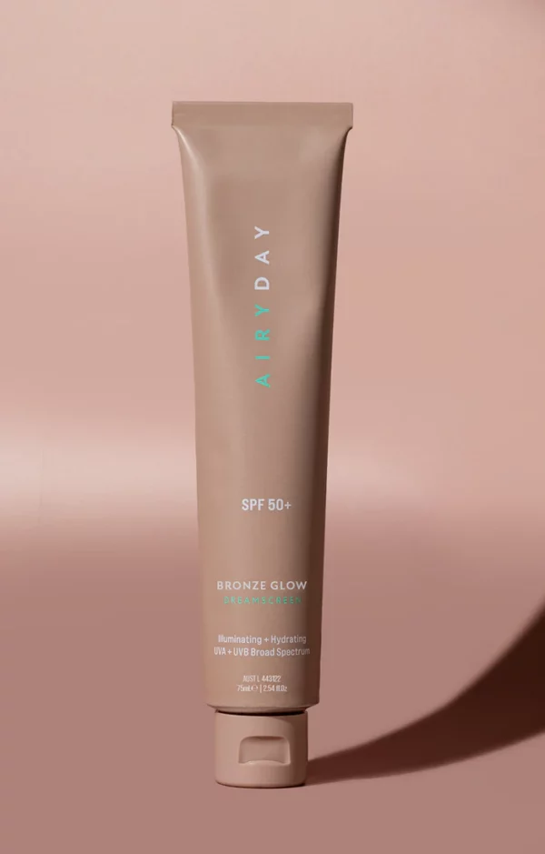 A tube of Golden Glow SPF50+ Dreamscreen 75ml stands upright on a matching beige background, casting a right-side shadow. The packaging displays product details in white and mint green text.