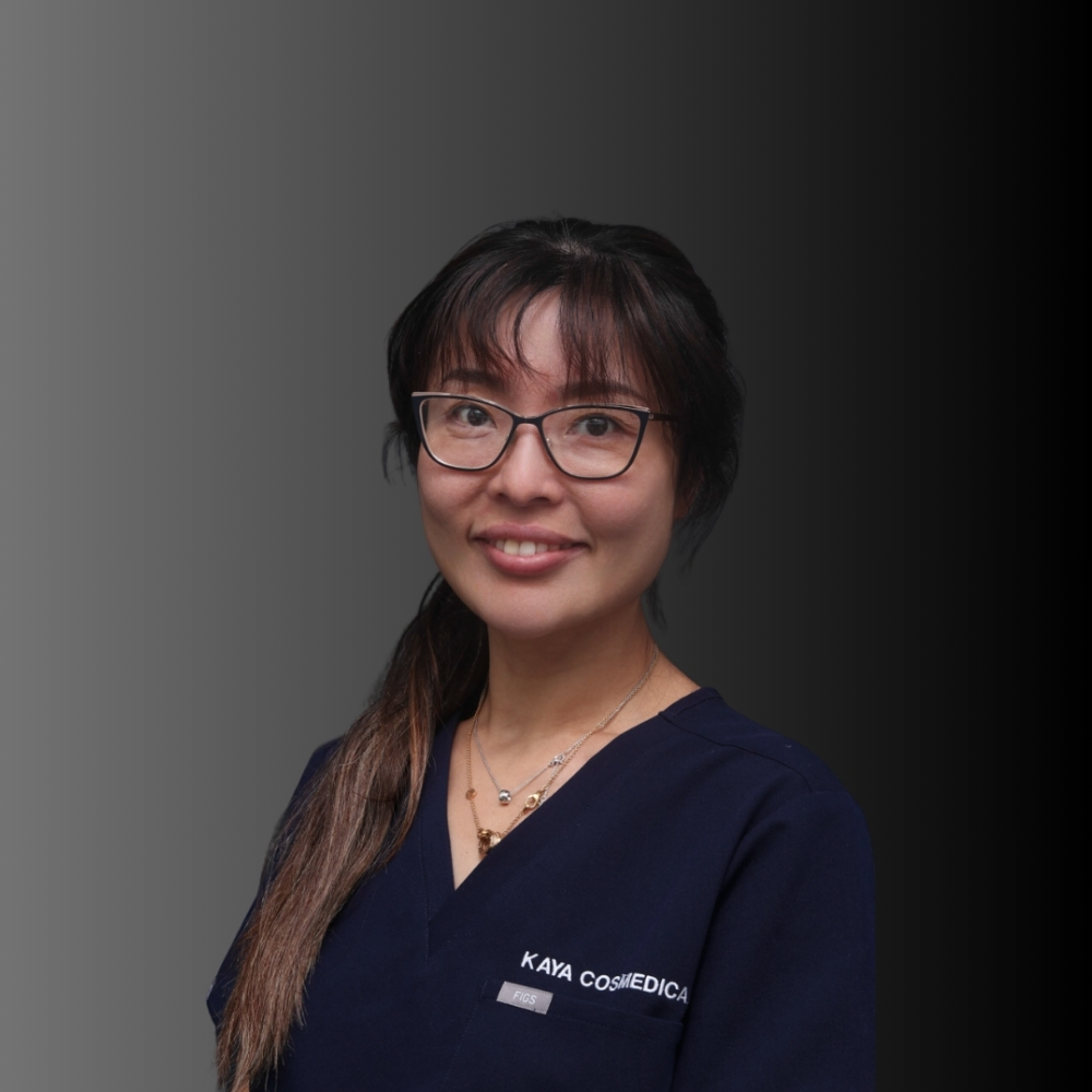 Doreen-2 - Kaya Cosmedica A woman with long dark hair, glasses, and wearing navy blue scrubs with "KAYA COSMEDICA" on the chest stands in front of a gradient black background, smiling at the camera.
