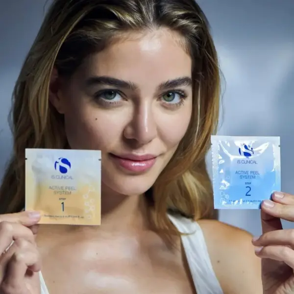 A woman with long hair holds two Is Clinical Active Peel System packets labeled "1" and "2" against a neutral background. The Is Clinical Active Peel System is also available in a convenient pack of 15.