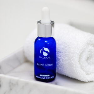 A blue glass dropper bottle labeled “Active Serum 15ml” stands upright on a white marble tray beside a rolled white towel.