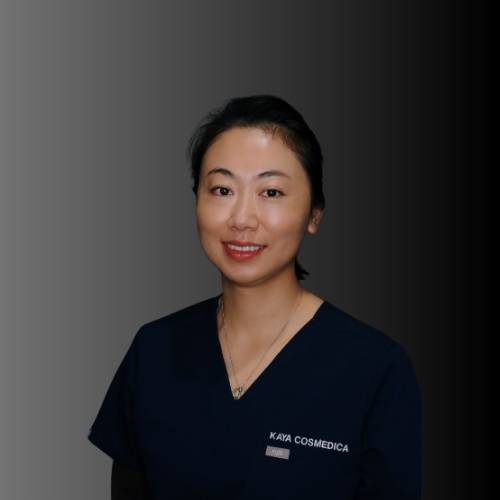 Janina - Kaya Cosmedica A woman wearing a navy blue scrub top with "KAYA COSMEDICA" written on it, smiling at the camera, standing against a gradient black and gray background.