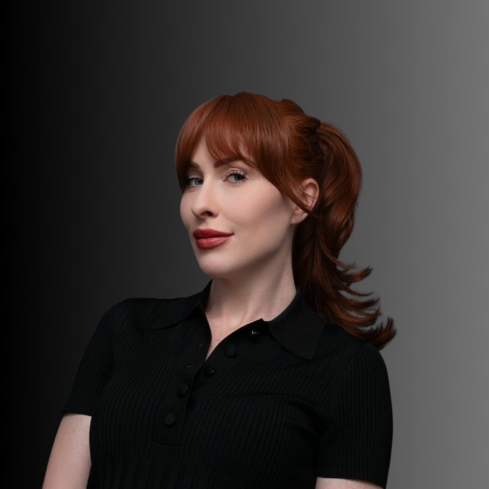 Juliette-2 - Kaya Cosmedica A woman with fair skin and long red hair styled in a loose ponytail is wearing a black short-sleeve collared shirt. She is posing in front of a dark-to-light gradient background.