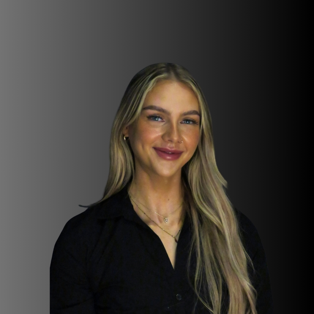 A woman with long blonde hair wearing a black shirt and layered necklaces stands in front of a gradient grey to black background, smiling at the camera.