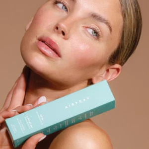 A woman with slicked-back hair and glowing skin holds a Fragrance Free - Clear as Day SPF50+ Dreamscreen 75ml box near her neck, gazing slightly upward against a neutral brown background.