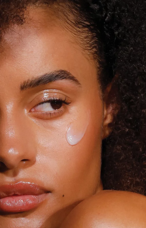 Close-up of a woman's face with curly hair and glossy makeup, featuring a dab of Clear as Day SPF50+ Dreamscreen 75ml on her cheekbone as she looks slightly to the side.