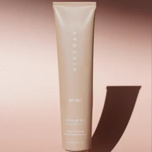 A beige tube labeled "Clear as Day SPF50+ Dreamscreen 75ml" stands upright on a light brown surface, casting a shadow to the right. The packaging notes its deep hydration and broad-spectrum UVA/UVB protection.