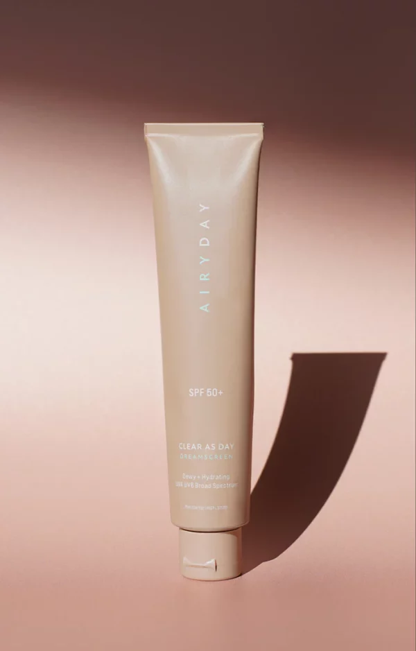 A beige tube labeled "Clear as Day SPF50+ Dreamscreen 75ml" stands upright on a light brown surface, casting a shadow to the right. The packaging notes its deep hydration and broad-spectrum UVA/UVB protection.