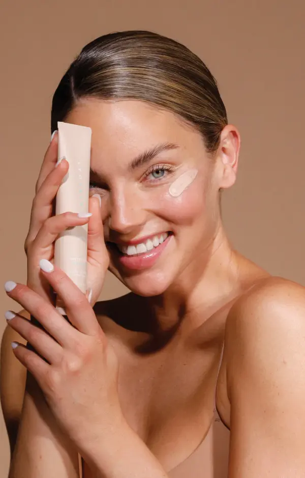 A woman with slicked-back hair smiles at the camera, holding a tube of Pretty in Zinc SPF50+ Dreamscreen 75ml beside her face. She has cream under one eye, wears a nude top, and poses against a beige background.