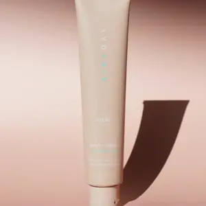 A beige tube of Pretty in Zinc SPF50+ Dreamscreen 75ml stands upright on a pink surface, casting a long shadow. The packaging features minimalist white and pale blue text.