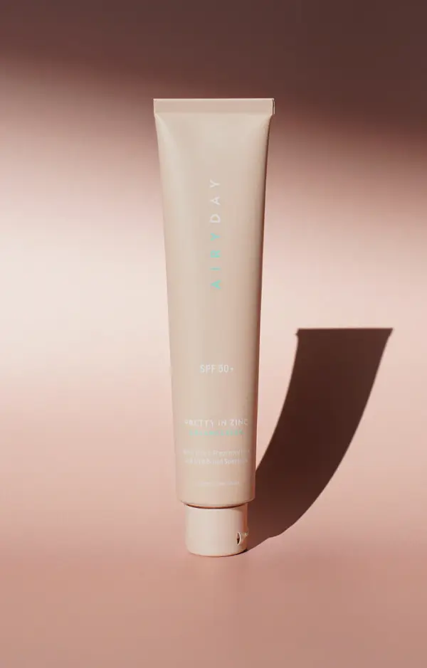A beige tube of Pretty in Zinc SPF50+ Dreamscreen 75ml stands upright on a pink surface, casting a long shadow. The packaging features minimalist white and pale blue text.