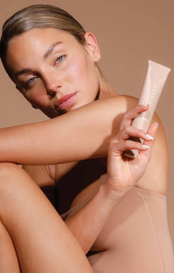 A woman with slicked-back hair holds the Fragrance Free - Clear as Day SPF50+ Dreamscreen 75ml, a visible patch of cream on her cheek. She poses in a nude-toned outfit against a neutral background, embodying the fragrance-free routine.