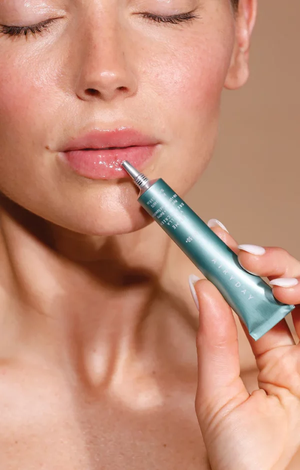 A fair-skinned person holds a metallic green tube of Lip Me Vanilla SPF50+ Dreamscreen 15ml lip balm near their slightly parted lips, eyes closed, showing glossy, sun-protected lips.