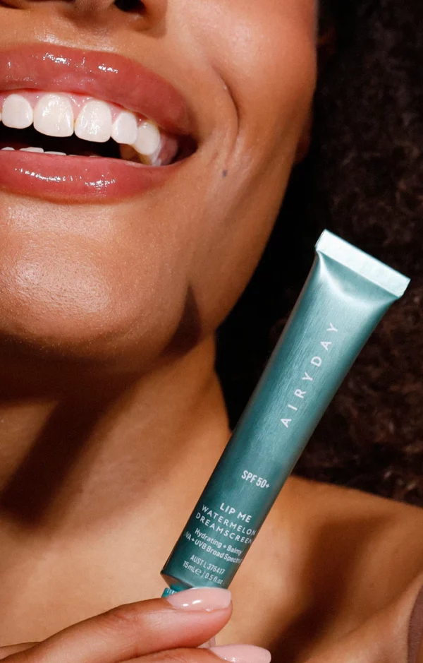 A person smiles, revealing white teeth, and holds a green tube of Lip Me Watermelon SPF50+ Dreamscreen 15ml lip balm labeled "SPF 50+" and "WATERMELON," with ingredients and product details visible.