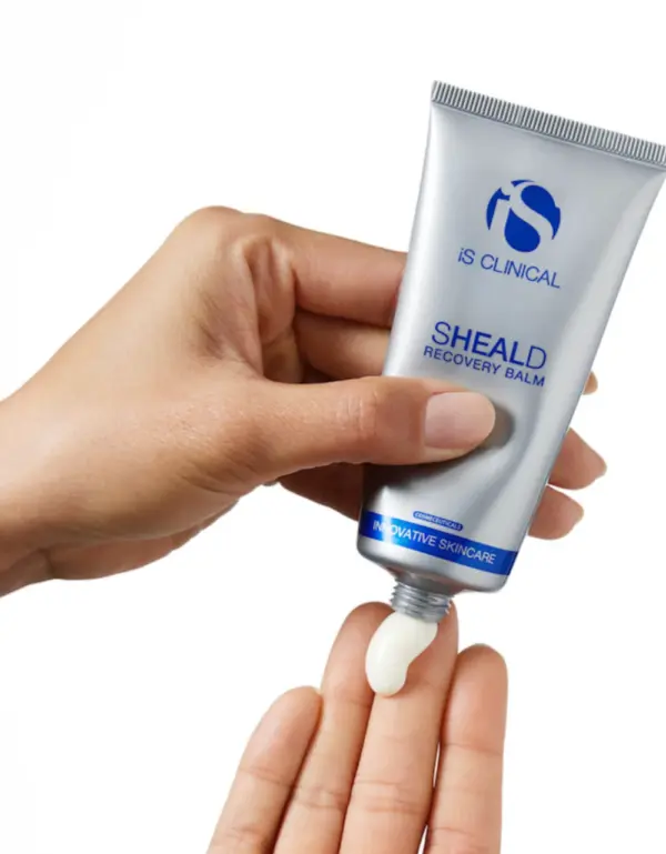 A person squeezes a small amount of Sheald Recovery Balm 15g, from a silver and blue tube, onto their fingertip against a white background.