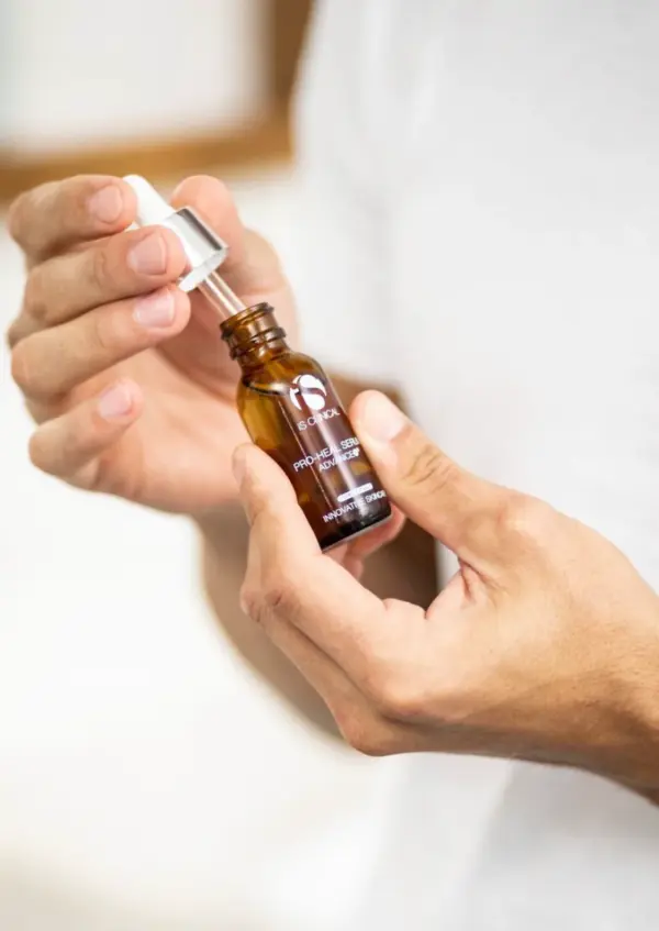 A hand holds an amber dropper bottle labeled “Pro-Heal Serum Advance+ 30ml,” with one hand unscrewing the cap. The label describes a serum for hair, while the background stays softly out of focus.